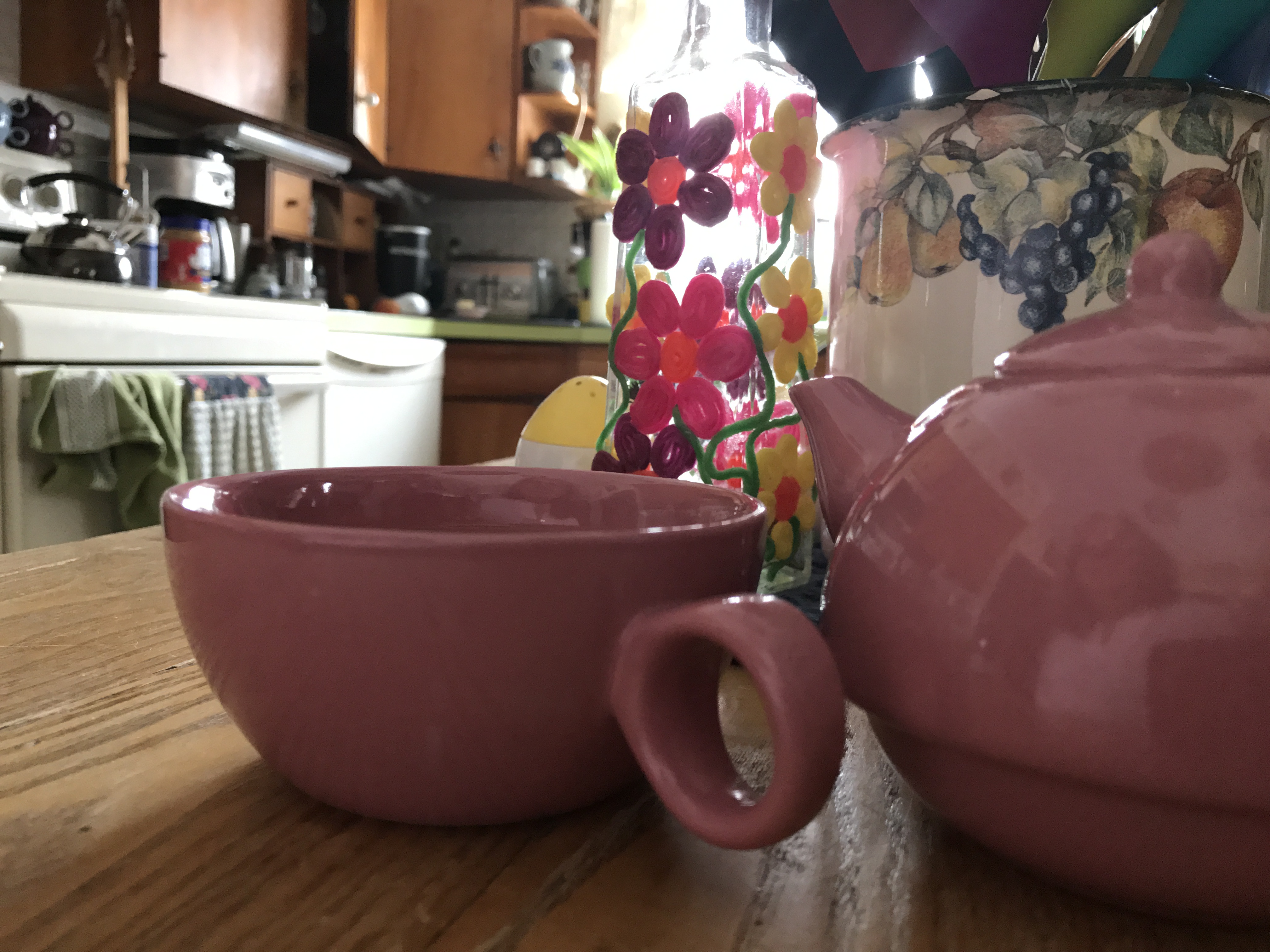 In a kitchen at the table where teacup and teapot are visible with a reflection of the photographer in the teapot