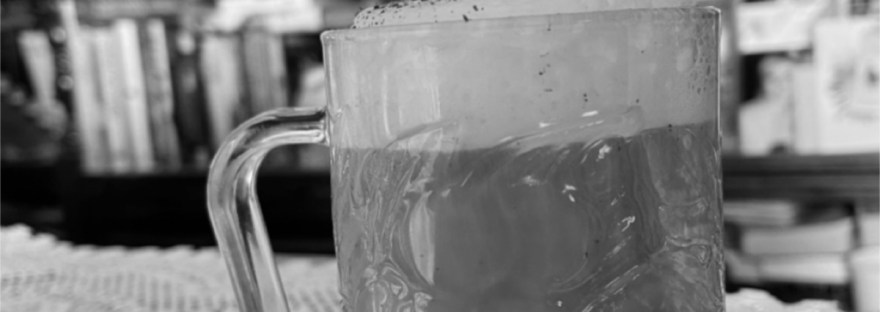 a frothed coffee in a clear mug on a doily in front of a filled bookcase