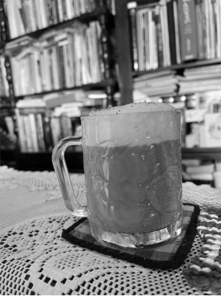a frothed coffee in a clear mug on a doily in front of a filled bookcase
