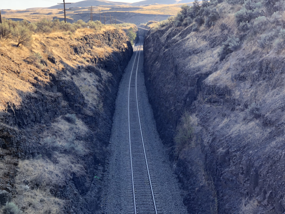 Railroad tracks in basalt cut below semi-arid landscape of sagebrush with miles of power lines along ancient lava flow landscape carved by less ancient massive glacial floods