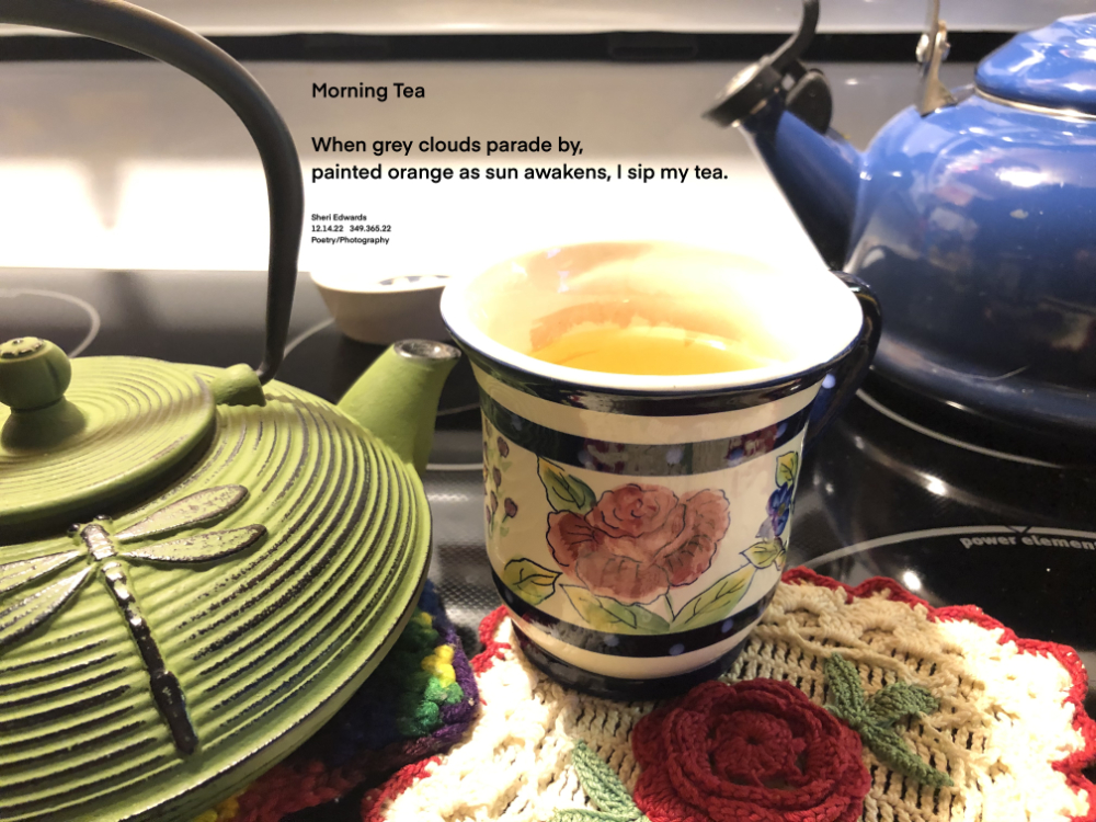 A green teapot with dragonfly, a multi-colored, flowery mug of tea, and a blue whistling teapot in a row near a crocheted rose potholder