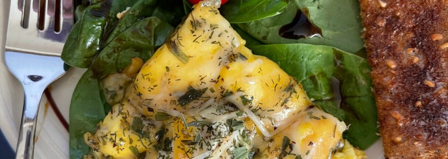 The Face of Breakfast: a triangle egg frittata with a cherry tomato at the tip on a bed of balsam vinegar topped spinach with two oranges at the top left/right edges of the plate and a side of whole grain toast