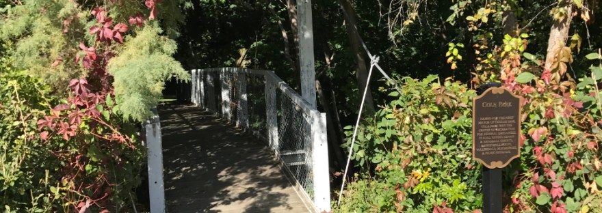 poem on sidewalk entrance to foliage-covered footbridge over Fiddle Creek leading to Cole Park.