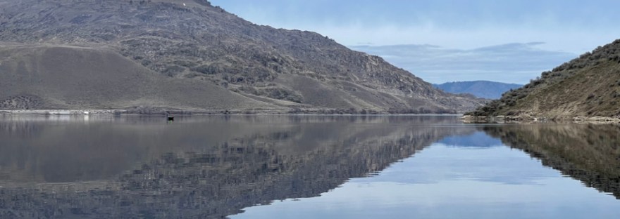 small boat and horizon reflected on Lake Roosevelt on Feb 3rd, 2024, a calm and silent, windless afternoon in the blue-grey hues of sky and lake